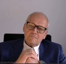 Älterer Mann mit Brille und blauem Anzug sitzt nachdenklich mit verschränkten Händen vor grauem Hintergrund.