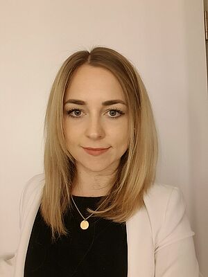 Porträt einer jungen Frau mit blonden, schulterlangen Haaren, die einen weißen Blazer über einem schwarzen Oberteil trägt.