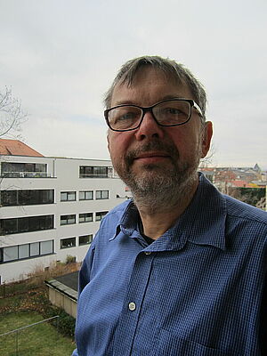 Mann mit Brille und blauem Hemd vor einem Gebäude und einer Stadtlandschaft im Hintergrund.