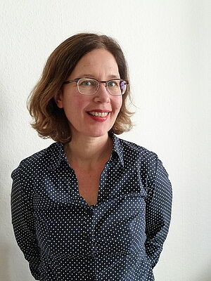 Frau mit kurzen braunen Haaren und Brille, die ein dunkelblaues, weiß gepunktetes Hemd trägt und vor einer weißen Wand steht.