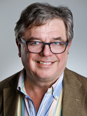 Lächelnder Mann mittleren Alters mit grauem Haar, Brille und braunem Sakko vor grauem Hintergrund.