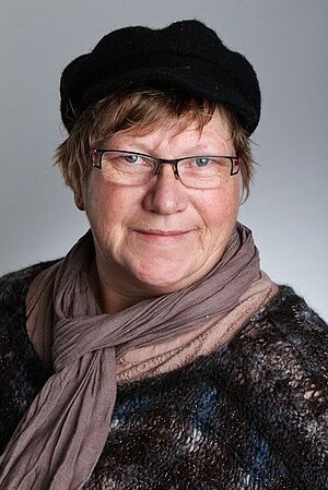 Ältere Frau mit Brille, schwarzem Hut und braunem Schal, leicht lächelnd vor grauem Hintergrund.