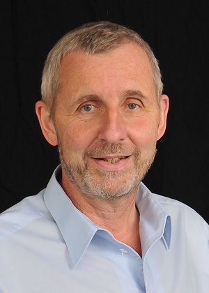 Älterer Mann mit kurzem grauem Haar, Bart und hellblauem Hemd vor schwarzem Hintergrund.