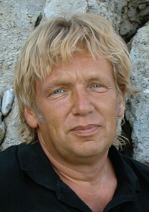 Porträt eines Mannes mit blonden Haaren und blauen Augen vor einem felsigen Hintergrund.