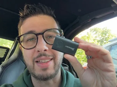 Man with glasses holding a small black AAWireless device inside a car.