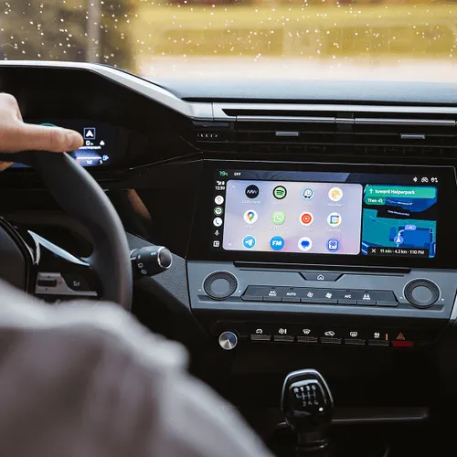 Driver's view of a car dashboard showing a multifunctional touchscreen with navigation and app icons, and a hand on the steering wheel.