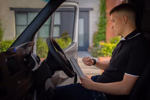 Man sitting in a vehicle reading a document while checking his watch, with an open door revealing a residential background.