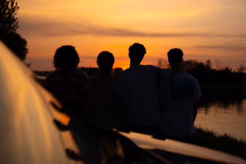 Four people standing by a calm river, silhouetted against a colorful sunset sky.