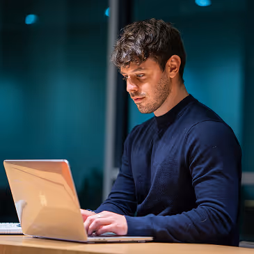 Man in navy sweater working on a laptop at a desk in a dimly lit room.