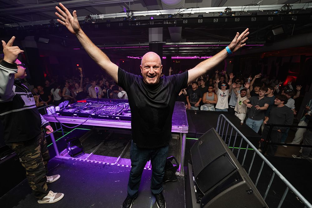 Marc Rousso DJing at a club. Marc has his arms high in the air, with a crowd of clubbers behind him enjoying the music.