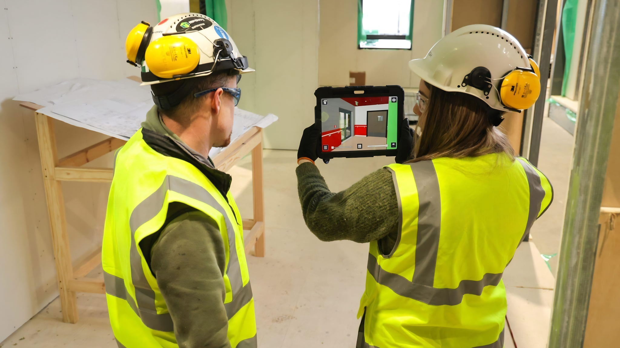 Two construction workers wearing high-visibility vests and hard hats on a building site, with one worker using a tablet to view interactive 3D digital work instructions and assembly models.