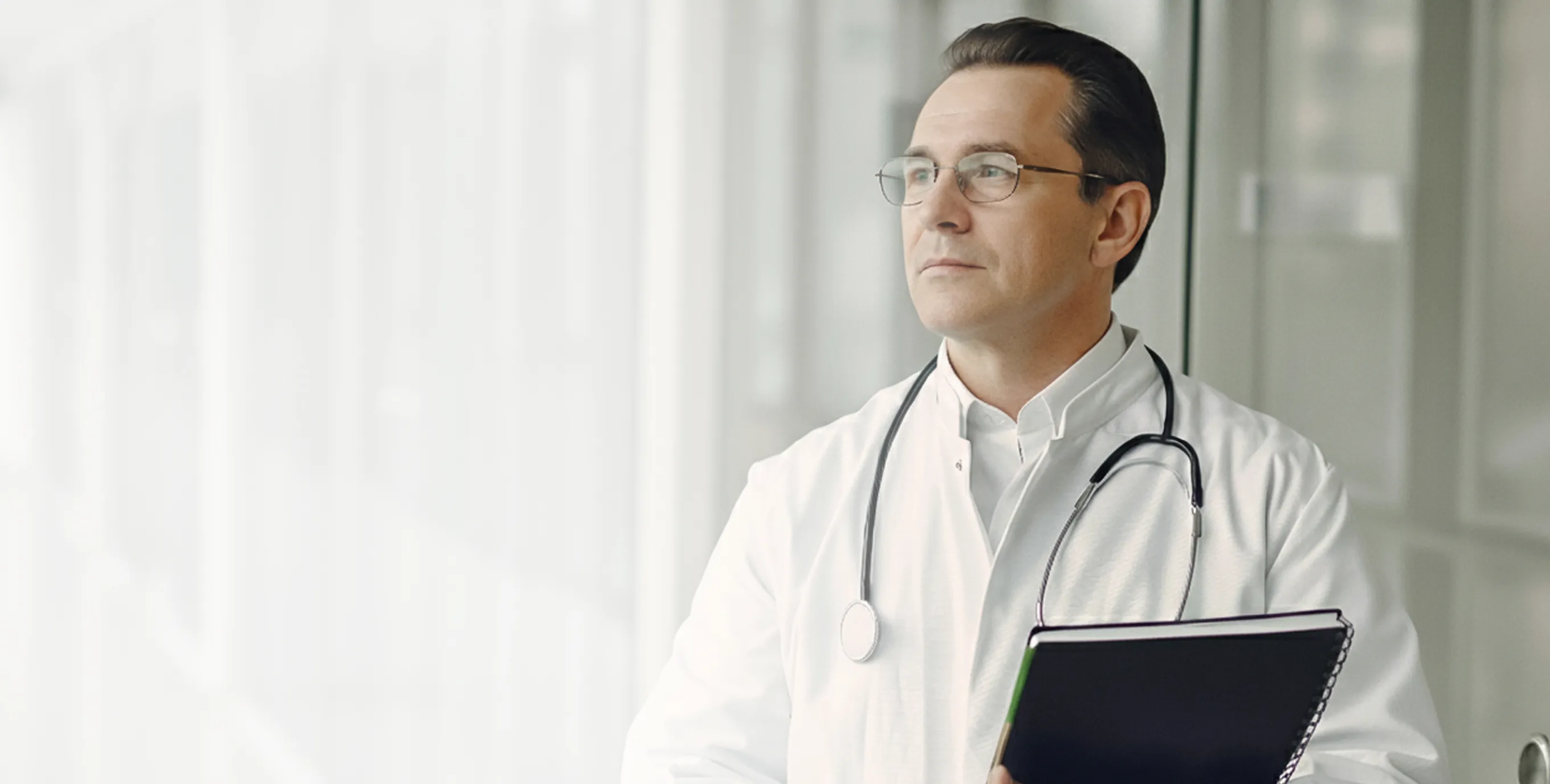Male doctor wearing glasses and a white coat with stethoscope, holding a black notebook and looking thoughtful.