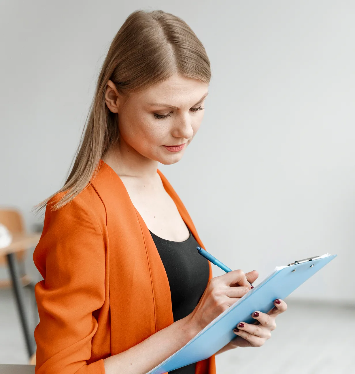 Woman in an orange blazer writing on a blue clipboard with a pen, looking focused.
