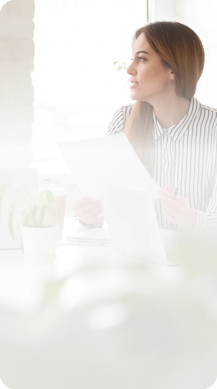 Young woman in a striped shirt holding papers and looking thoughtfully to the side at a bright workspace.