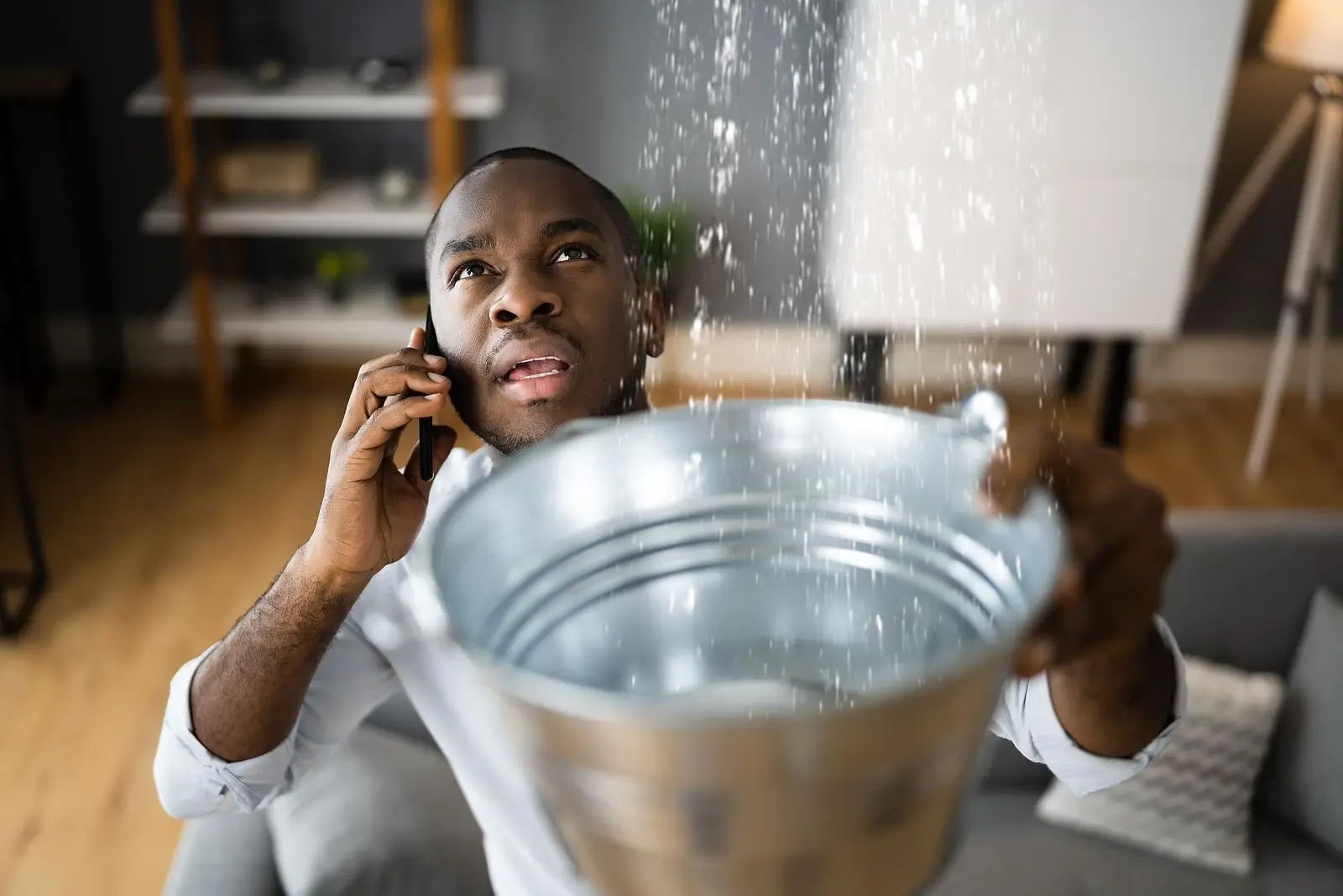 Black man holding a bucket under a leak while calling for a plumber