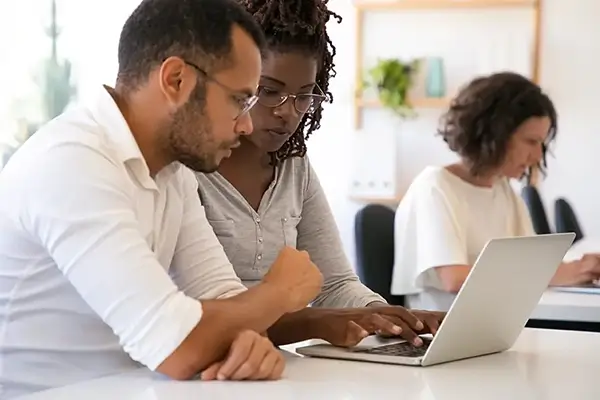 Man and women working together on a laptop in a room with other people