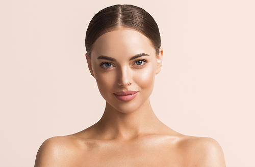 Woman with smooth, glowing skin and slicked-back hair smiling slightly while posing against a soft beige background, showcasing a healthy and radiant complexion.