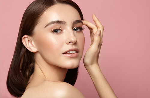 Woman with glowing skin and straight brown hair posing with one hand near her eyebrow, looking confidently at the camera against a soft pink background.