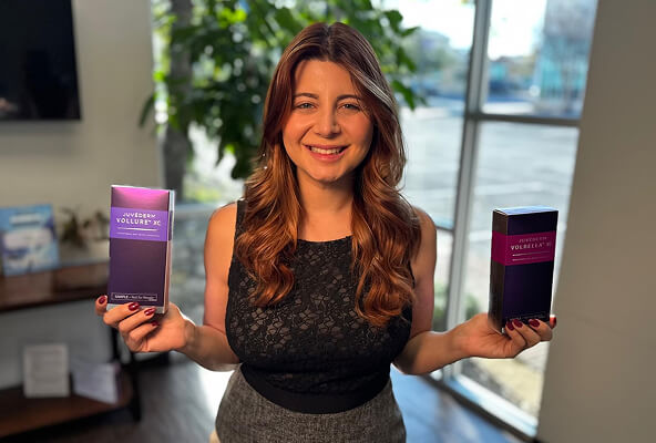 Jessica Aldakar, smiling and holding boxes of Juvéderm Vollure and Volbella, stands in a well-lit room with large windows and greenery, representing her role at Reveal Medical Aesthetics.