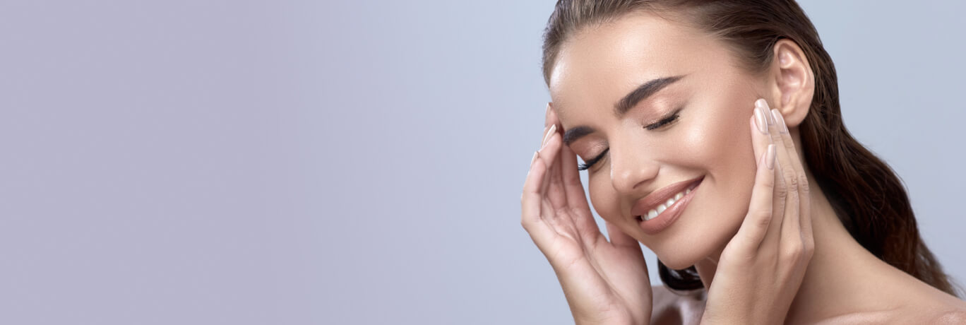 Smiling woman with glowing skin and closed eyes gently touching her face with both hands, radiating confidence and beauty against a soft gradient background.