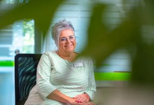 Ältere Frau mit grauen kurzen Haaren und Brille, lächelnd und sitzend in einem modernen Raum, teilweise hinter grünen Blättern sichtbar.