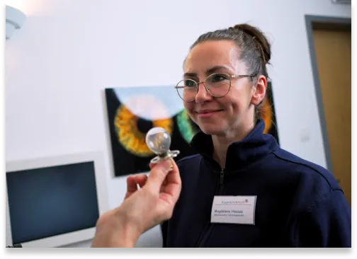 Frau mit Brille und dunkler Arbeitsjacke lächelt, während ihr jemand ein kleines Objekt vorhält.
