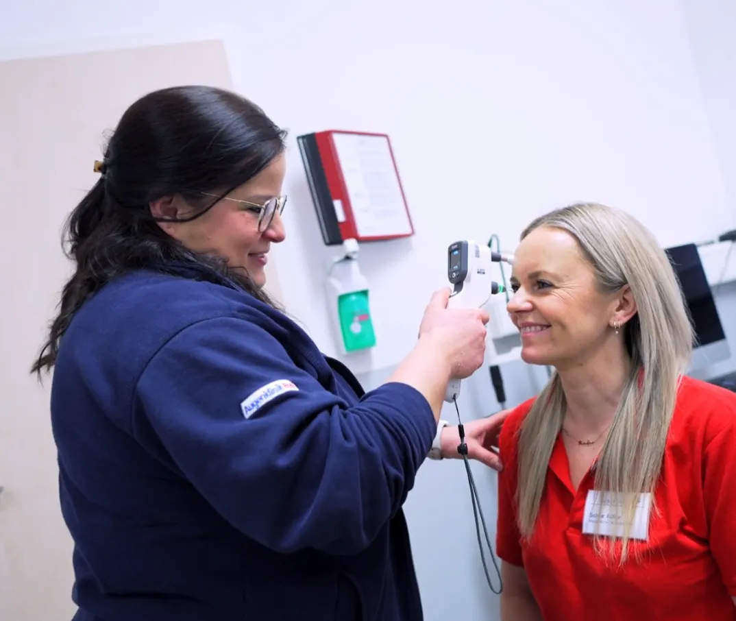 Eine Frau misst mit einem Ohrthermometer die Temperatur einer lächelnden Frau in einem roten Hemd.