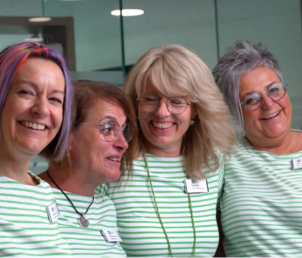 Vier lächelnde Frauen in grünen und weißen gestreiften T-Shirts stehen zusammen in einem modernen Raum.