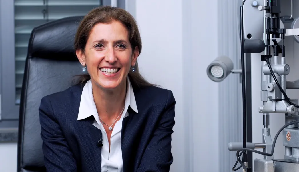 Lächelnde Frau in Business-Kleidung sitzt vor einem Augenuntersuchungsgerät in einem Untersuchungszimmer.