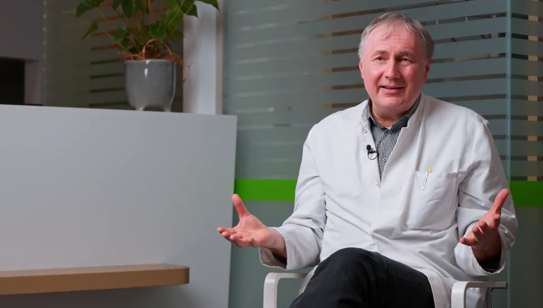 Ein älterer Mann in weißem Arztkittel sitzt gestikulierend auf einem Stuhl in einem modernen Büro.