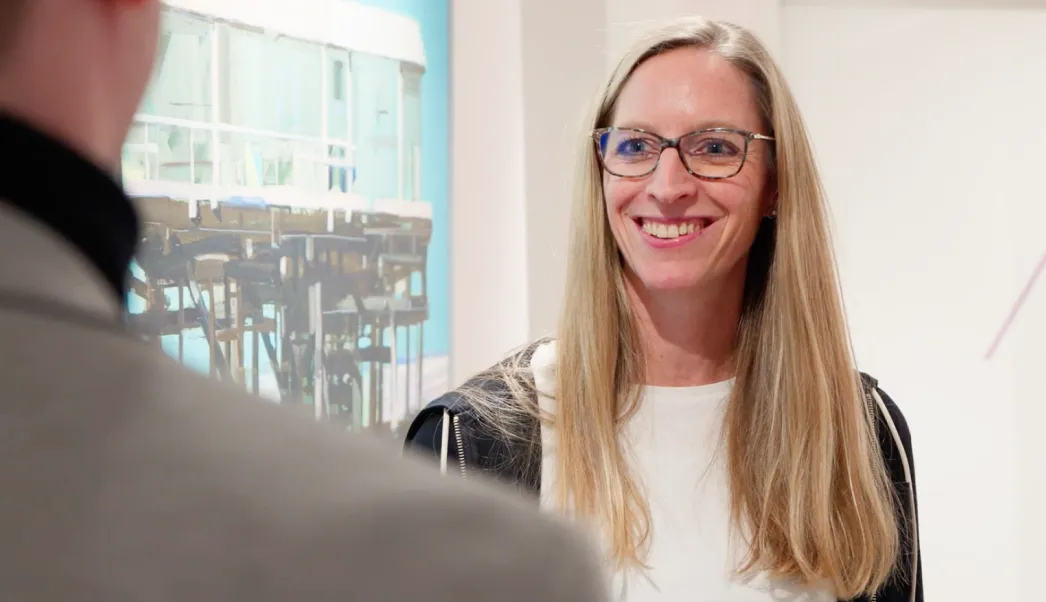 Frau mit langen blonden Haaren und Brille lächelt bei einem Gespräch mit einer anderen Person.