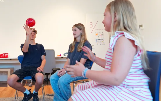 Drei Jugendliche sitzen auf Stühlen in einem Raum, ein Junge wirft einen roten Ball, während zwei Mädchen zuschauen.
