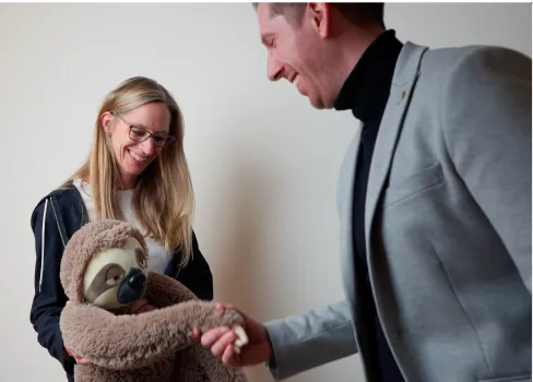 Eine lächelnde Frau hält eine Stofftierfaultierpuppe, während ein Mann ihr die Hand schüttelt.