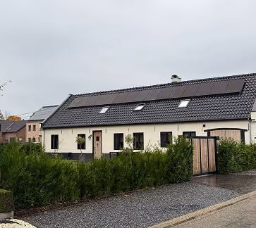 Witte woning met een zwart dak bekleed met zonnepanelen, omgeven door groene hagen en een oprit van grind.