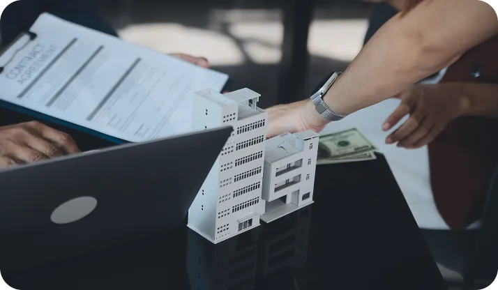 Person holding a contract agreement near a laptop and miniature building models on a table, with another person extending their hand holding cash.