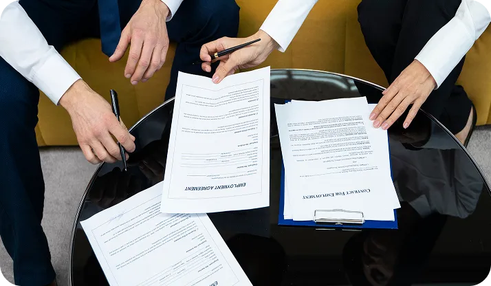 Two people sitting at a round glass table reviewing and signing employment agreement documents.