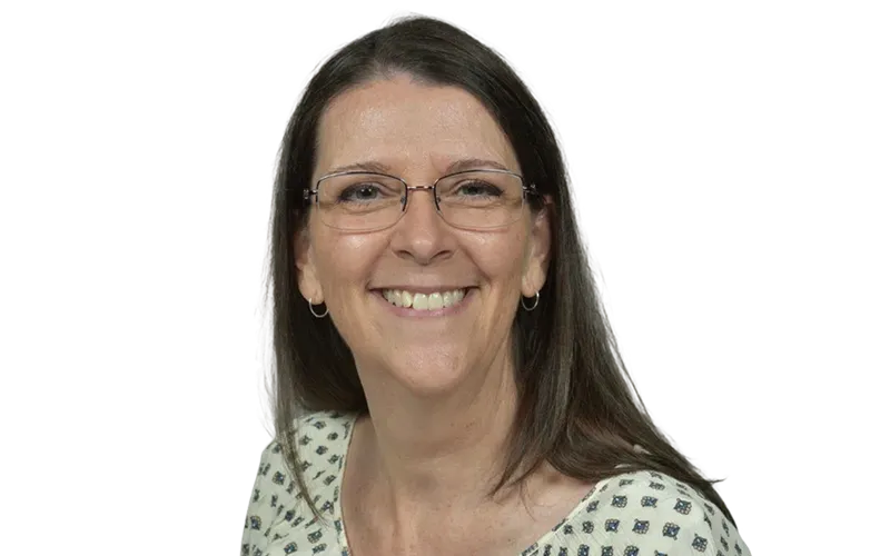 Smiling woman with long brown hair, glasses, and hoop earrings wearing a patterned blouse.
