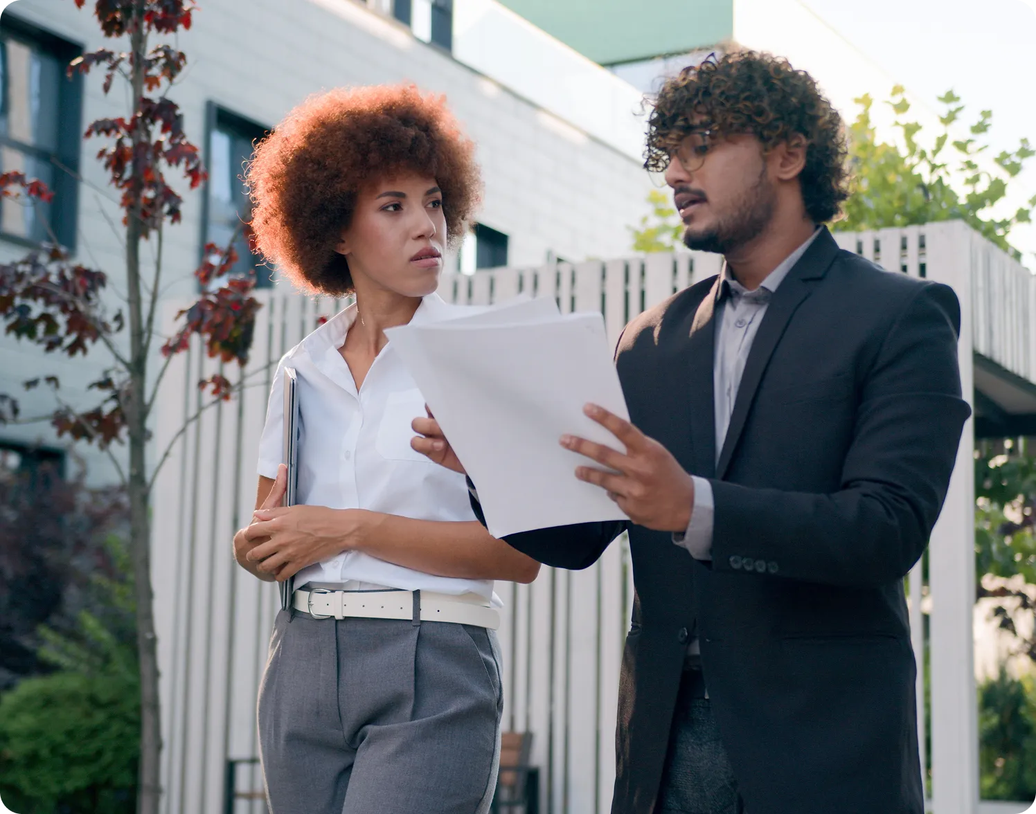 A man in a black blazer explains documents to a woman in a white shirt and gray pants outside an office building.