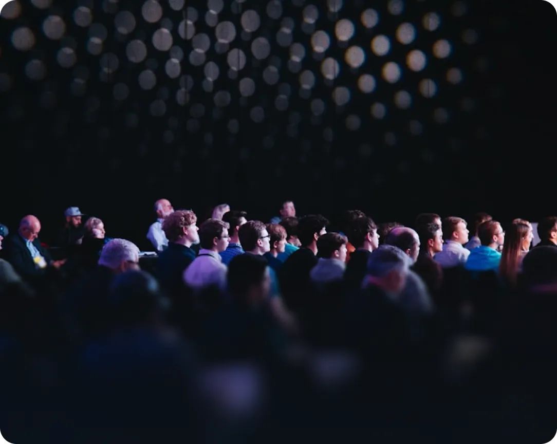 Audience of people seated and attentively watching a presentation or event in a dark room with circular light patterns on the background wall.