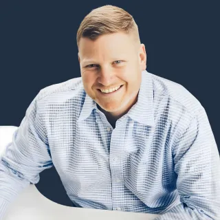Woman with blond hair wearing a brown plaid blazer and a gold necklace, looking directly at the camera.
