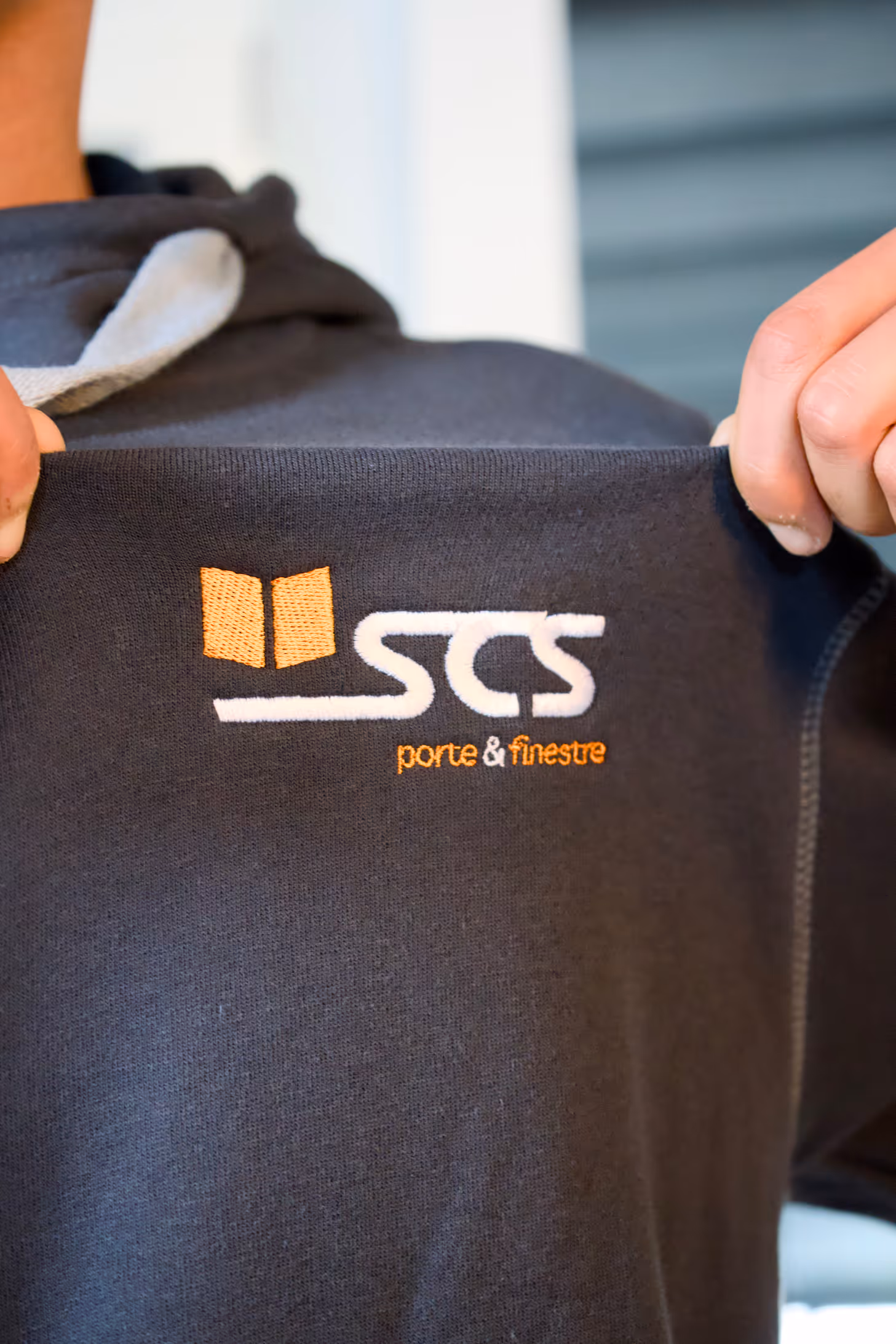 Person holding up a black garment featuring the embroidered logo 'SCS porte & finestre' in white and orange.
