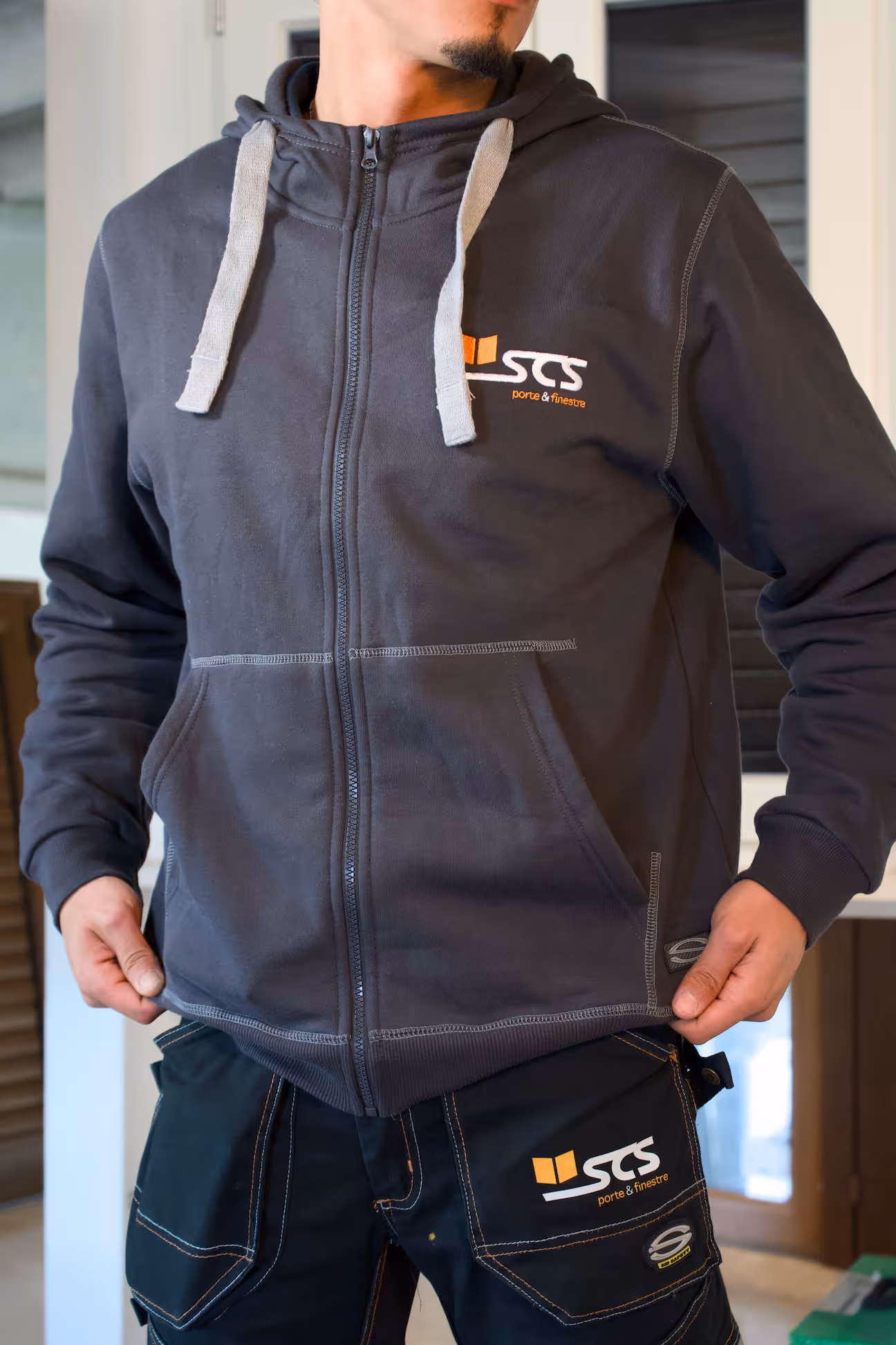 Person wearing a dark gray zip-up hoodie and work pants with the SCS porte & finestre logo on both garments.