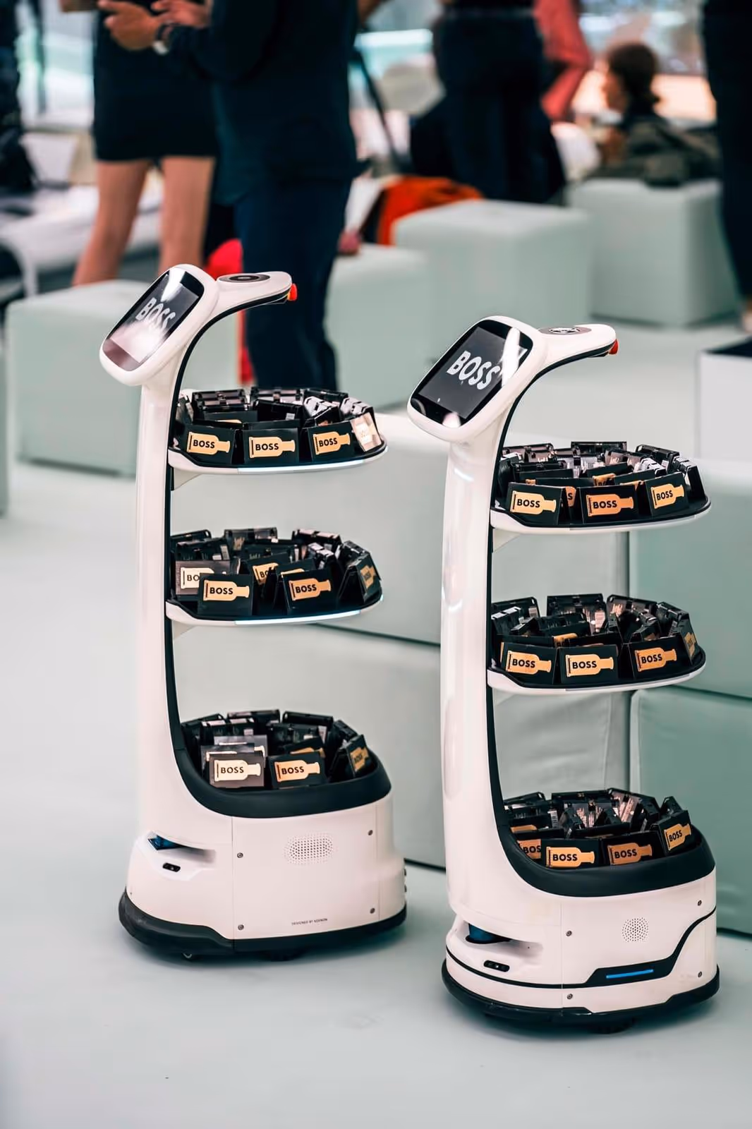 Two white service robots with screens labeled 'BOSS' holding trays of mini bottles with 'BOSS' labels at an indoor event.