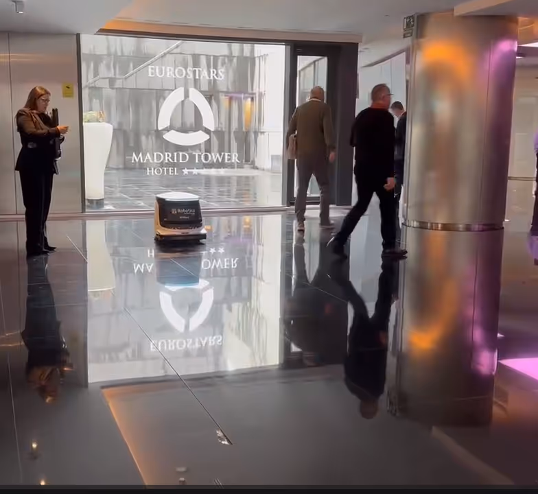 A small autonomous cleaning robot moving on the reflective floor of a hotel foyer with Eurostars Madrid Tower Hotel logo visible on the glass wall.