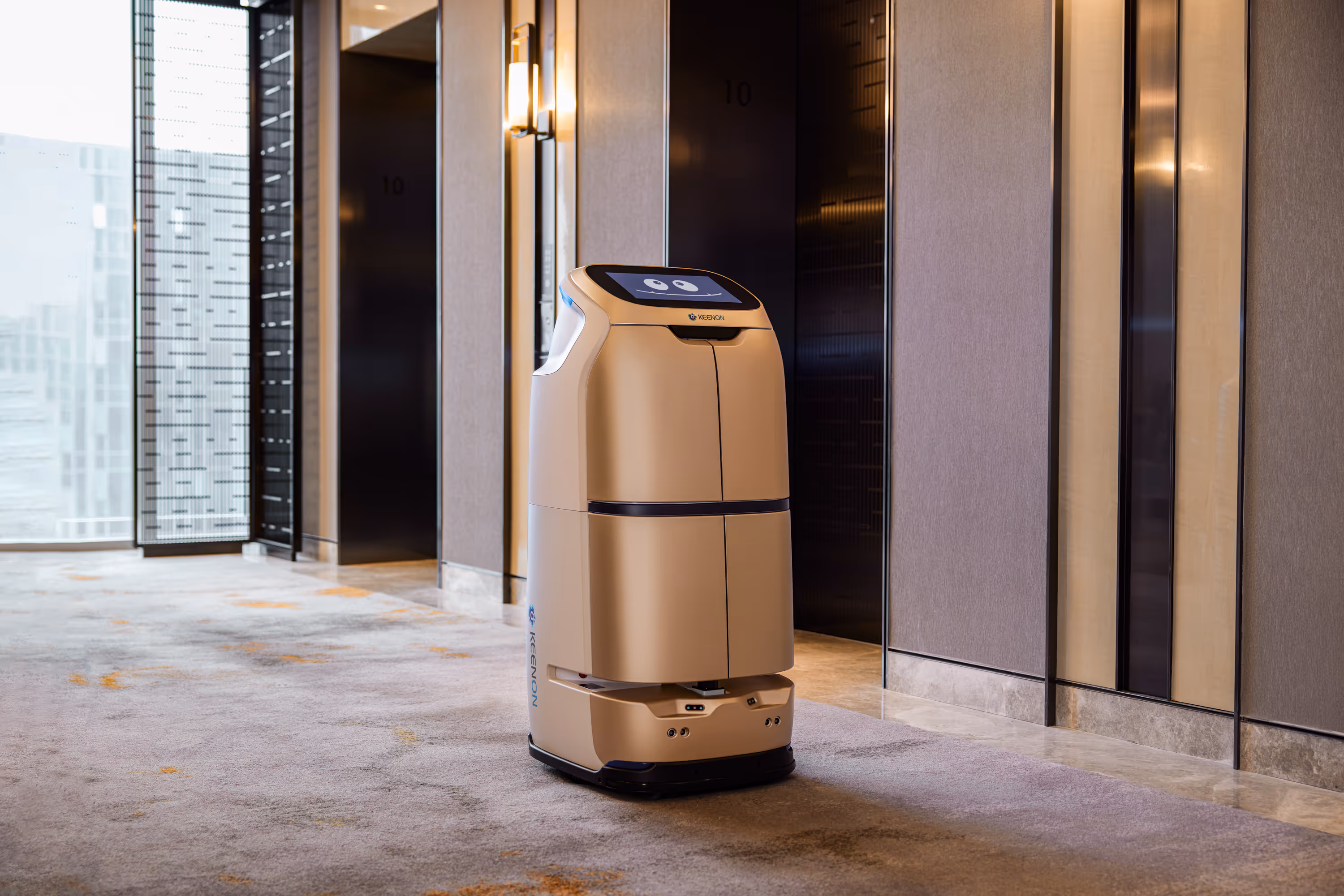 Gold-colored service robot with a smiling face screen standing in a modern building hallway near elevator doors.