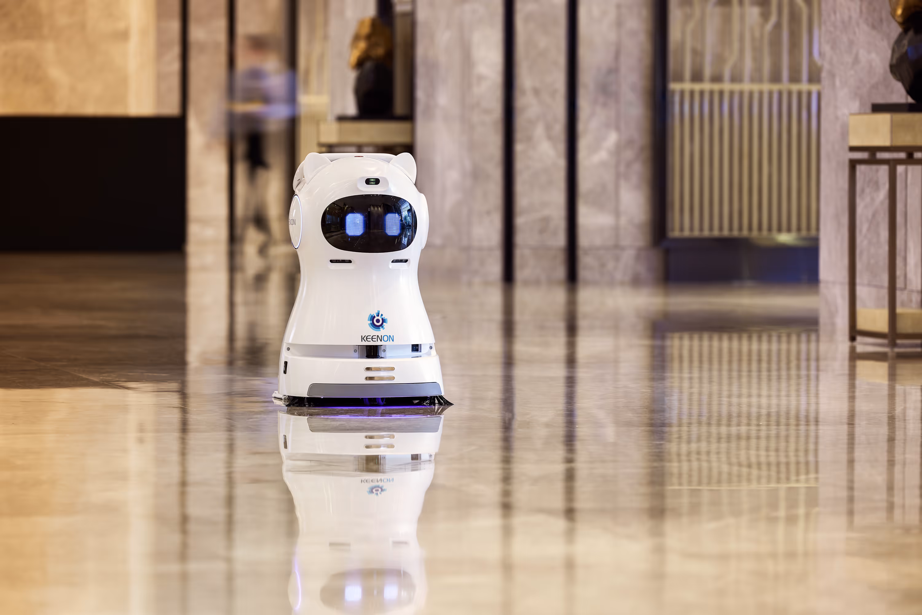 White Keenon cleaning robot with digital eyes operating on a shiny marble floor indoors.