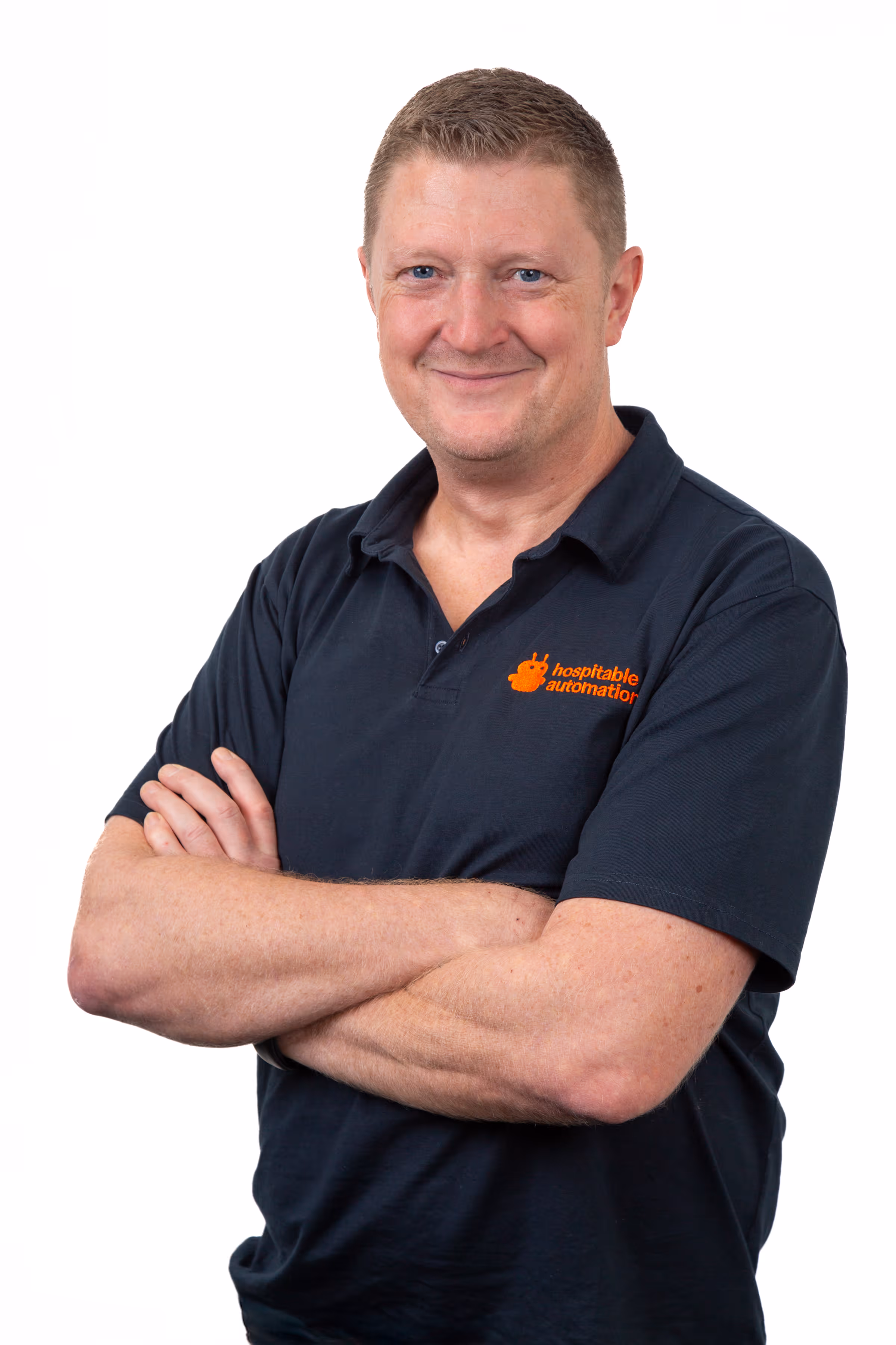Man wearing a black polo shirt with an orange 'hospitable automation' logo, smiling with arms crossed.