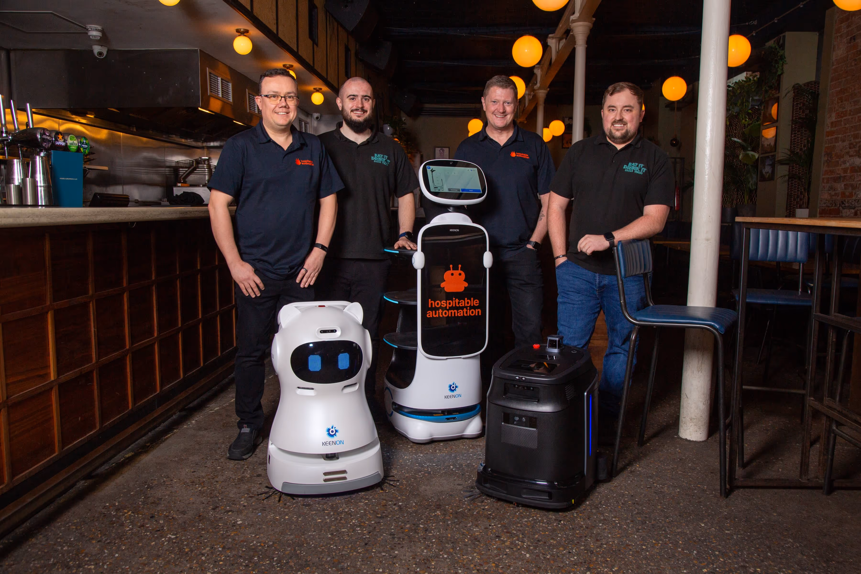 Four men standing inside a restaurant next to three service robots, one with a screen displaying 'hospitable automation'.