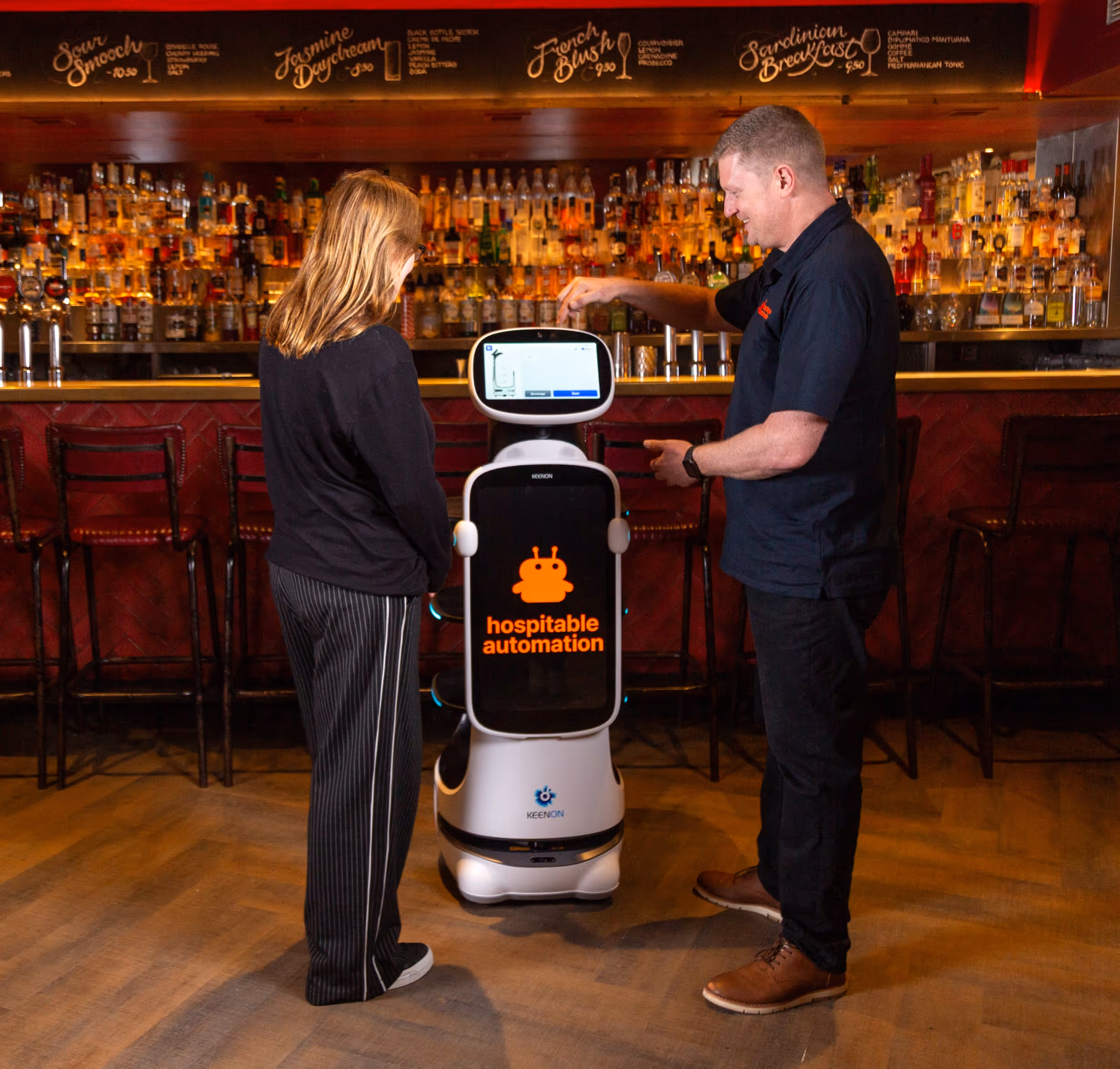 A man and a woman interact with a hospitality service robot in a bar with a well-stocked liquor shelf behind them.