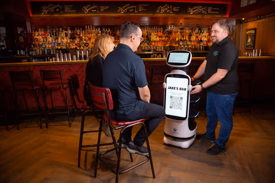 A bartender interacts with two seated customers at a bar using a service robot displaying a QR code for Jake's Bar.